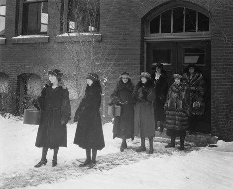 A group of women sets out from a brick building at Iowa State University in the winter of 1915. All are wearing coats and hats, and carying hatboxes. Several of the coats appear to be made of fur (racoon?); others appear to be a heavy wool. 